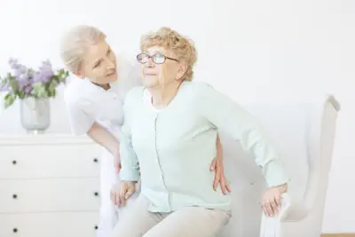 Young caregiver assisting smiling elderly lady in standing up from chair.